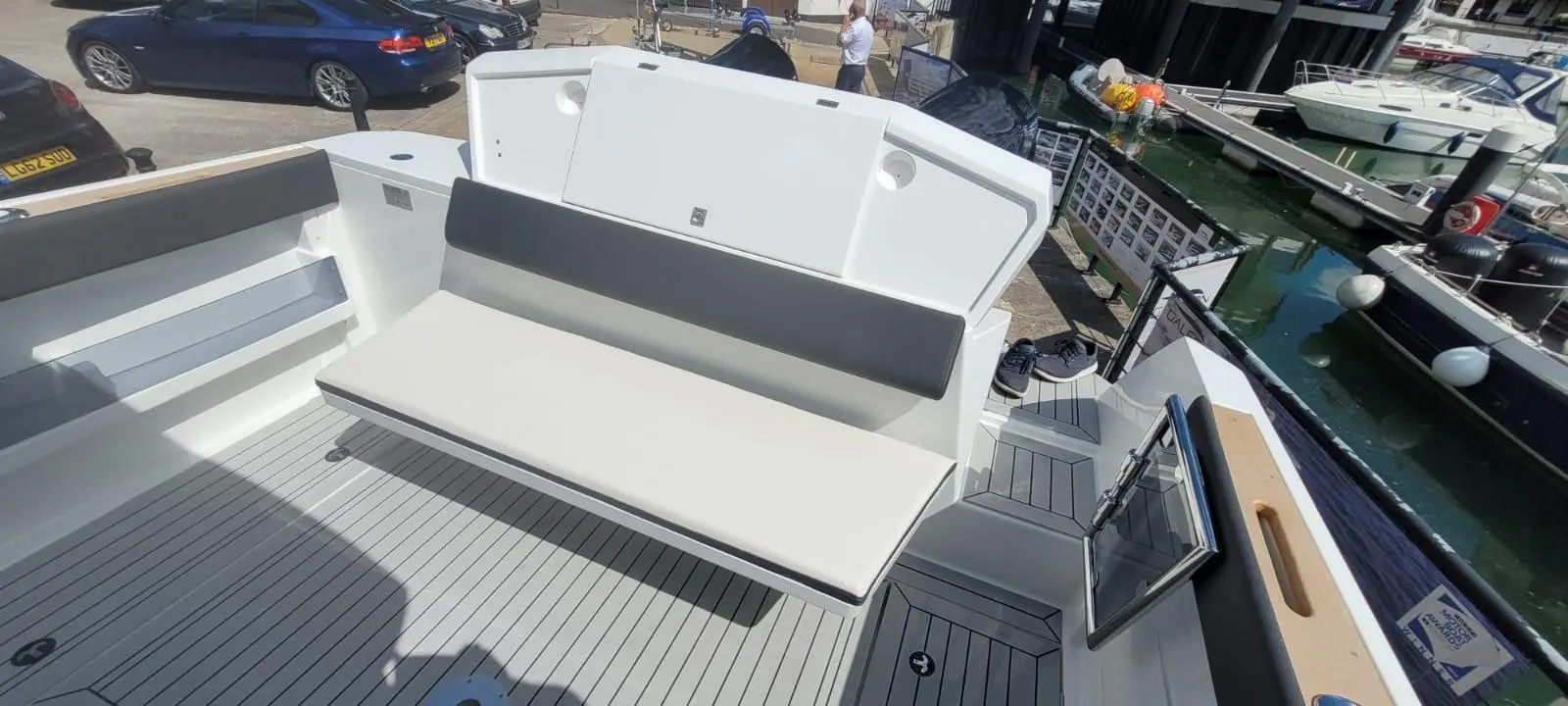 A sleek boat's deck features a grey cushioned bench, surrounded by marina boats and parked cars, under clear skies.